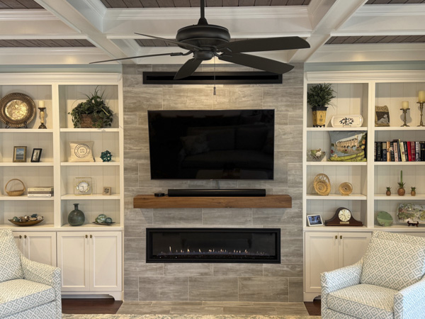 Custom fireplace wall in a Charleston SC living room — floor-to-ceiling stacked stone surround, floating live-edge wood mantel, linear gas fireplace with ember bed, and flanking white built-in cabinets with curated shelf decor by KDC Interiors