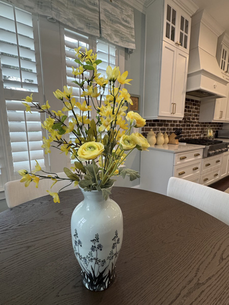 Dining table styling detail in a Mount Pleasant SC home — yellow forsythia and ranunculus floral arrangement in a blue-and-white chinoiserie vase on an oak table, with white kitchen cabinetry and brick backsplash beyond by KDC Interiors