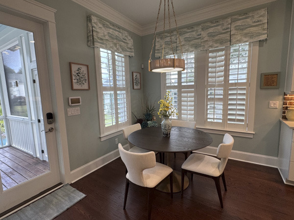 Light-filled breakfast nook in a Charleston SC home — round dark wood dining table with cream wingback chairs, brass-and-wood drum pendant, watercolor Roman shades over plantation shutters, and glass porch door by KDC Interiors
