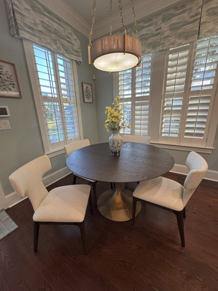 Elegant dining area in a Mount Pleasant SC residence — round pedestal table with brushed brass base, ivory wingback dining chairs on hardwood floors, illuminated drum chandelier, and layered window treatments by KDC Interiors