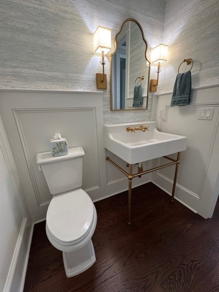 Elegant powder room in a Charleston SC home — white wainscoting with panel molding, textured silver grasscloth wallpaper, console sink with brushed brass legs and cross-handle faucet, gold-framed arched mirror, matching brass wall sconces with linen shades, and dark hardwood floors by KDC Interiors