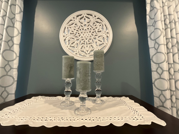 Curated bedroom vignette in a Charleston SC home — crystal candlestick holders with sage pillar candles, white eyelet lace runner, and carved medallion wall art against a moody teal accent wall by KDC Interiors