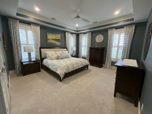 Spacious master bedroom retreat in Charleston SC — coffered tray ceiling with crown molding, plantation shutters with patterned linen drapes, dark wood bedroom furniture, and a coastal sunset painting by KDC Interiors