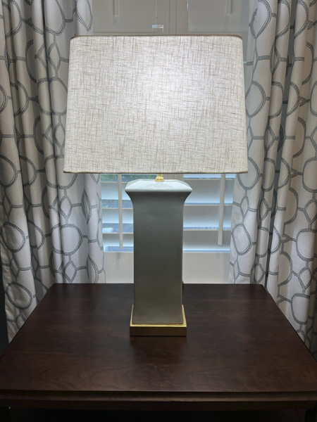 Designer bedside lighting in a Charleston SC bedroom — sage green ceramic table lamp with linen shade and brushed gold base on a dark wood nightstand, framed by geometric circle-pattern drapes selected by KDC Interiors