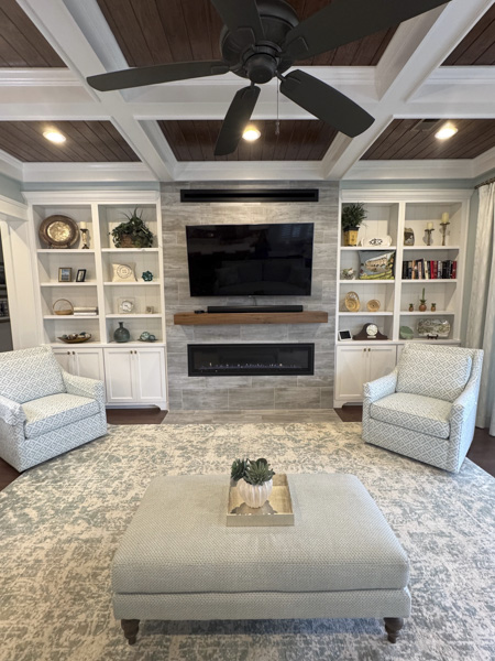 Transitional living room in a Charleston SC home — stained wood coffered ceiling, stacked stone linear fireplace with live-edge walnut mantel, custom white built-in bookshelves, and patterned swivel armchairs designed by KDC Interiors