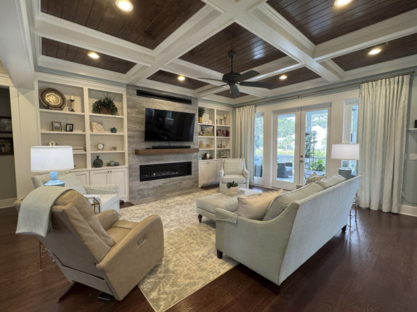 Open-concept family room in Mount Pleasant SC — dark wood coffered ceiling with recessed lighting, stone fireplace wall with floating mantel, neutral linen sofa and recliner, and French doors to a screened porch by KDC Interiors