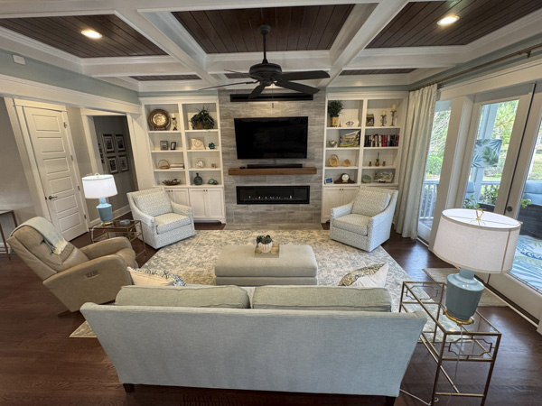 Coastal Charleston SC living room with full interior design — wood-paneled coffered ceiling, stone media wall with linear gas fireplace, styled built-in shelving, gold-and-glass side tables, and layered neutral textiles by KDC Interiors