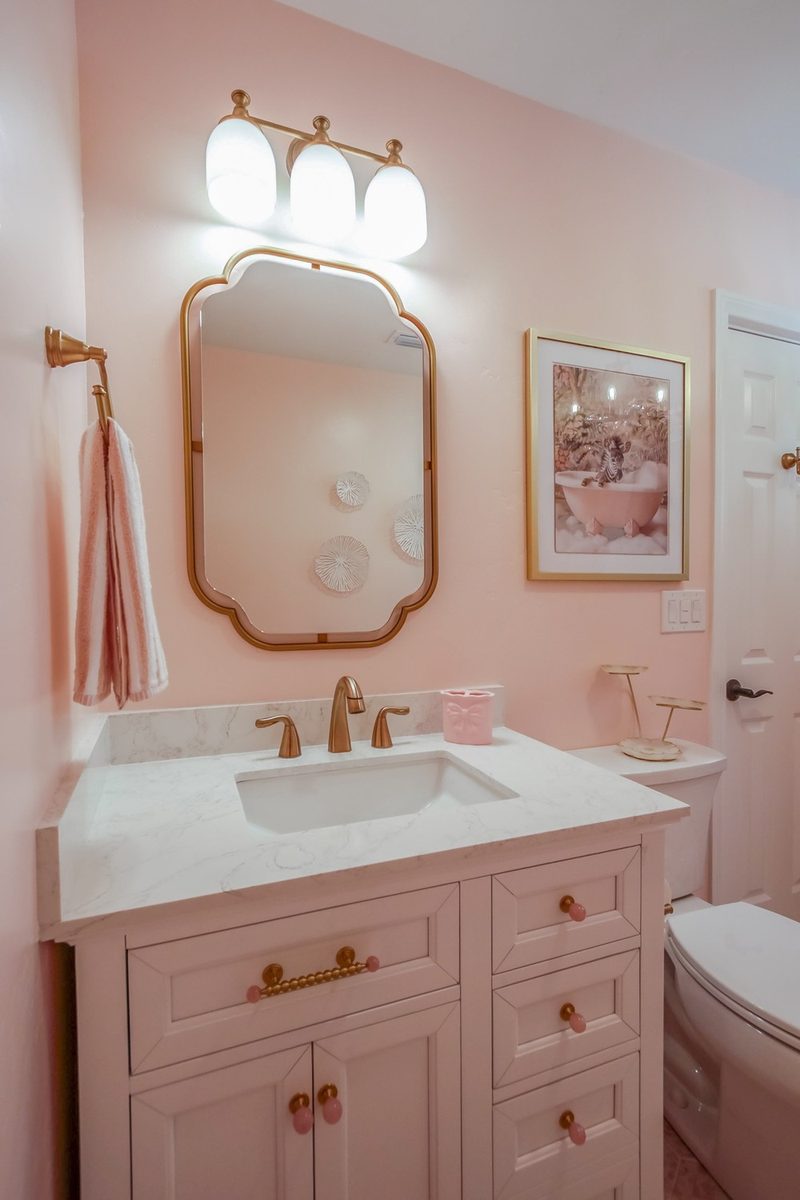 Pink vanity with a gold scalloped mirror, brass faucet, and brass and pink quartz hardware, by KDC Interiors
