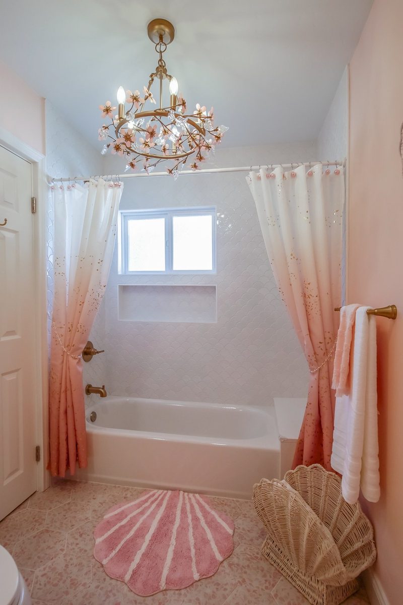 Tub beneath the whimsical floral chandelier with a pink shell rug and scalloped shower tile, by KDC Interiors