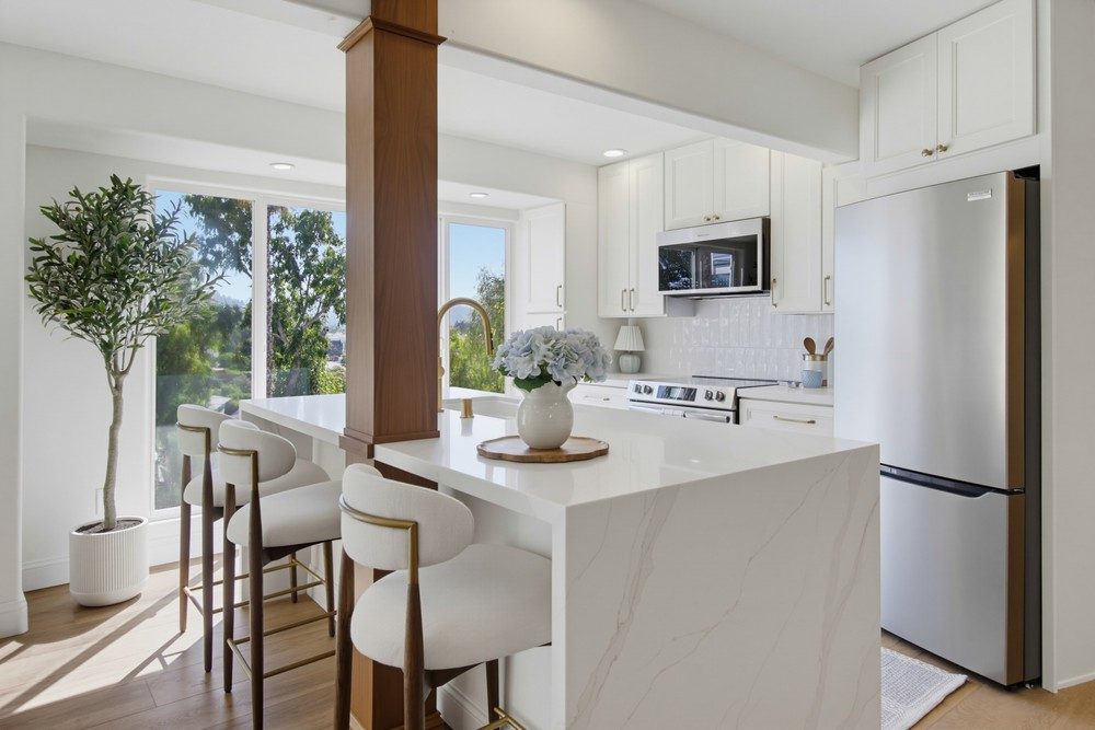 After: the open kitchen with white shaker cabinets, a waterfall quartz island, structural support post, and an olive tree at the window, by KDC Interiors