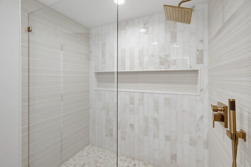 Walk-in shower with vertical marble tile and a brass rain head, by KDC Interiors